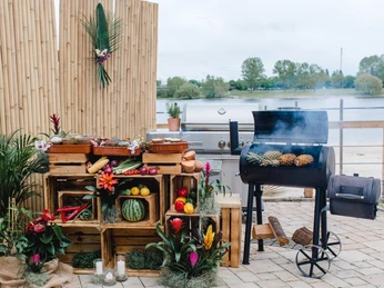 BBQ Grillplatz am See mit Holzkohlegrill, Obst, Gemüse und Holzkisten vor Schilfwand, dekoriert mit Blumen.Barbecue area by the lake with charcoal grill, fruit, vegetables and wooden crates in front of a reed wall, decorated with flowers.