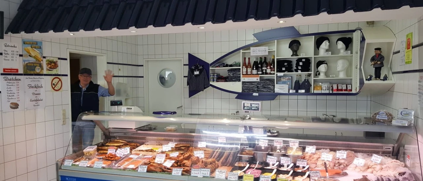 Allräucherei Prahm-Theke-Apen.jpg Ein Verkäufer in weißem Kittel grüßt freundlich hinter einer gut bestückten Fischtheke.