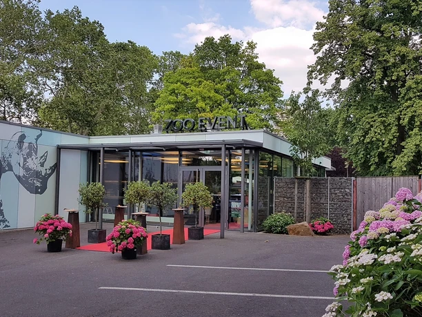 Zoo Event Eingang eines modernen Eventgebäudes mit Glasfront, flankiert von bunten Blumenarrangements.</p>