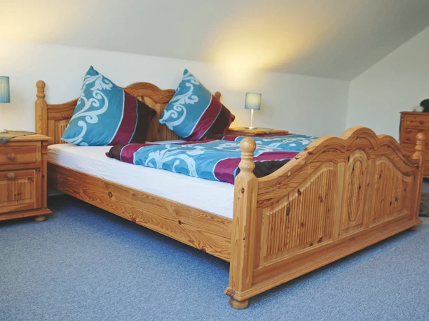 FEWO_Enie_3_DoppelzimmerCC-BY-SA_Teutoburger_Wald_Stadt_Schloss_Holte-Stukenbrock.jpg Ein geräumiges Doppelzimmer mit einem holzgeschnitzten Bett, farbiger Bettwäsche und Nachttischen.