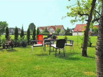 Garten mit Tisch, Stühlen und Sträuchern, umgeben von Wiesen und Häusern im Hintergrund.