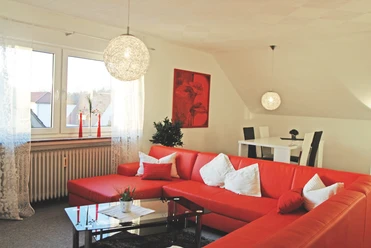 FEWO_Enie_1_WohnzimmerCC-BY-SA_Teutoburger_Wald_Stadt_Schloss_Holte-Stukenbrock.jpg Helles Wohnzimmer mit roten Sofas und weißem Esstisch, dekoriert mit Pflanzen und Kunst an der Wand.