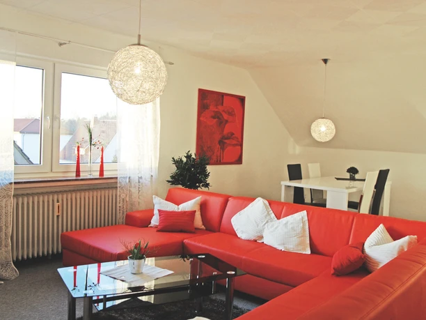 FEWO_Enie_1_WohnzimmerCC-BY-SA_Teutoburger_Wald_Stadt_Schloss_Holte-Stukenbrock.jpg Helles Wohnzimmer mit roten Sofas und weißem Esstisch, dekoriert mit Pflanzen und Kunst an der Wand.