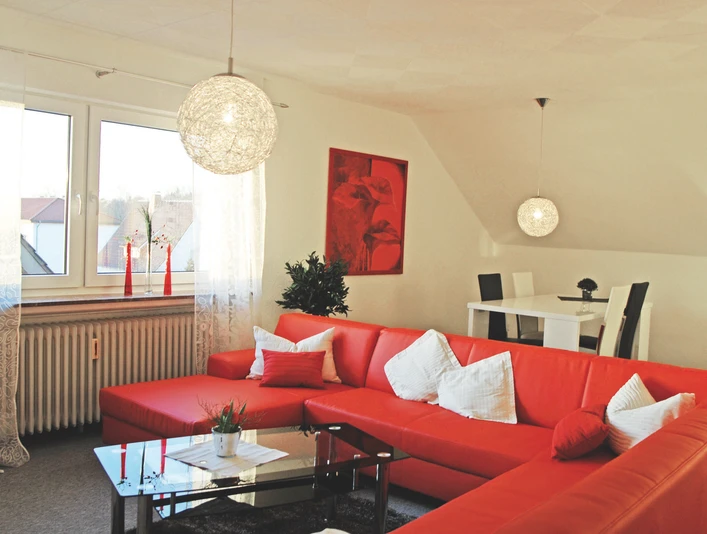 FEWO_Enie_1_WohnzimmerCC-BY-SA_Teutoburger_Wald_Stadt_Schloss_Holte-Stukenbrock.jpg Helles Wohnzimmer mit roten Sofas und weißem Esstisch, dekoriert mit Pflanzen und Kunst an der Wand.