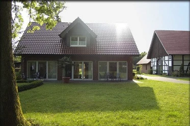 FeWo_Christa_Erichlandwehr.jpg Ein zweistöckiges Haus mit roten Dachziegeln, umgeben von gepflegtem Rasen und Bäumen bei Sonnenlicht.