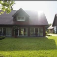 FeWo_Christa_Erichlandwehr.jpg Ein zweistöckiges Haus mit roten Dachziegeln, umgeben von gepflegtem Rasen und Bäumen bei Sonnenlicht.