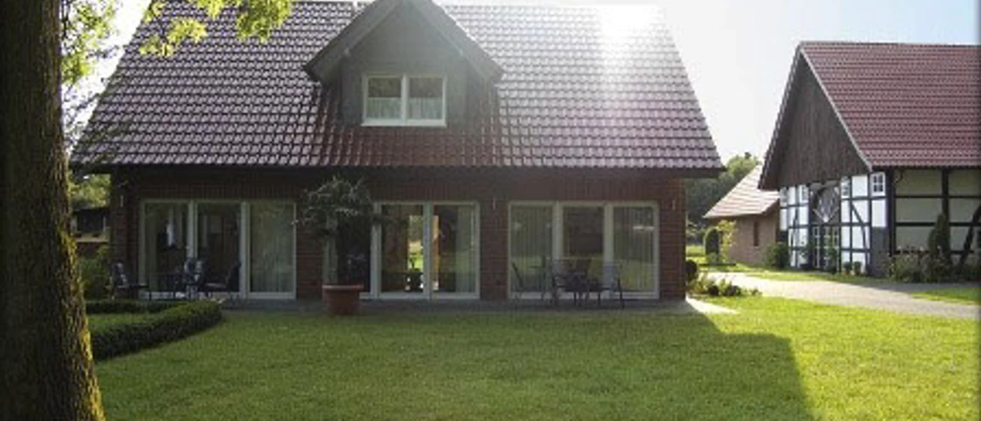 FeWo_Christa_Erichlandwehr.jpg Ein zweistöckiges Haus mit roten Dachziegeln, umgeben von gepflegtem Rasen und Bäumen bei Sonnenlicht.