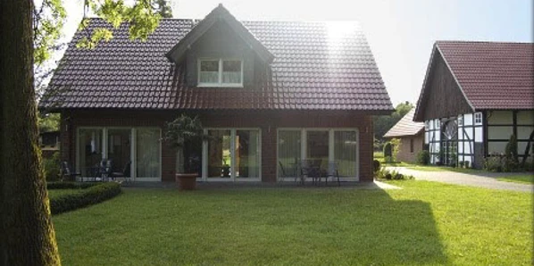 FeWo_Christa_Erichlandwehr.jpg Ein zweistöckiges Haus mit roten Dachziegeln, umgeben von gepflegtem Rasen und Bäumen bei Sonnenlicht.