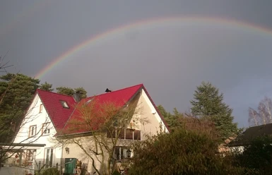 DSC_0232 Herzog.JPG Zweistöckiges Haus mit rotem Dach im Vordergrund, beeindruckender Regenbogen am Himmel darüber.