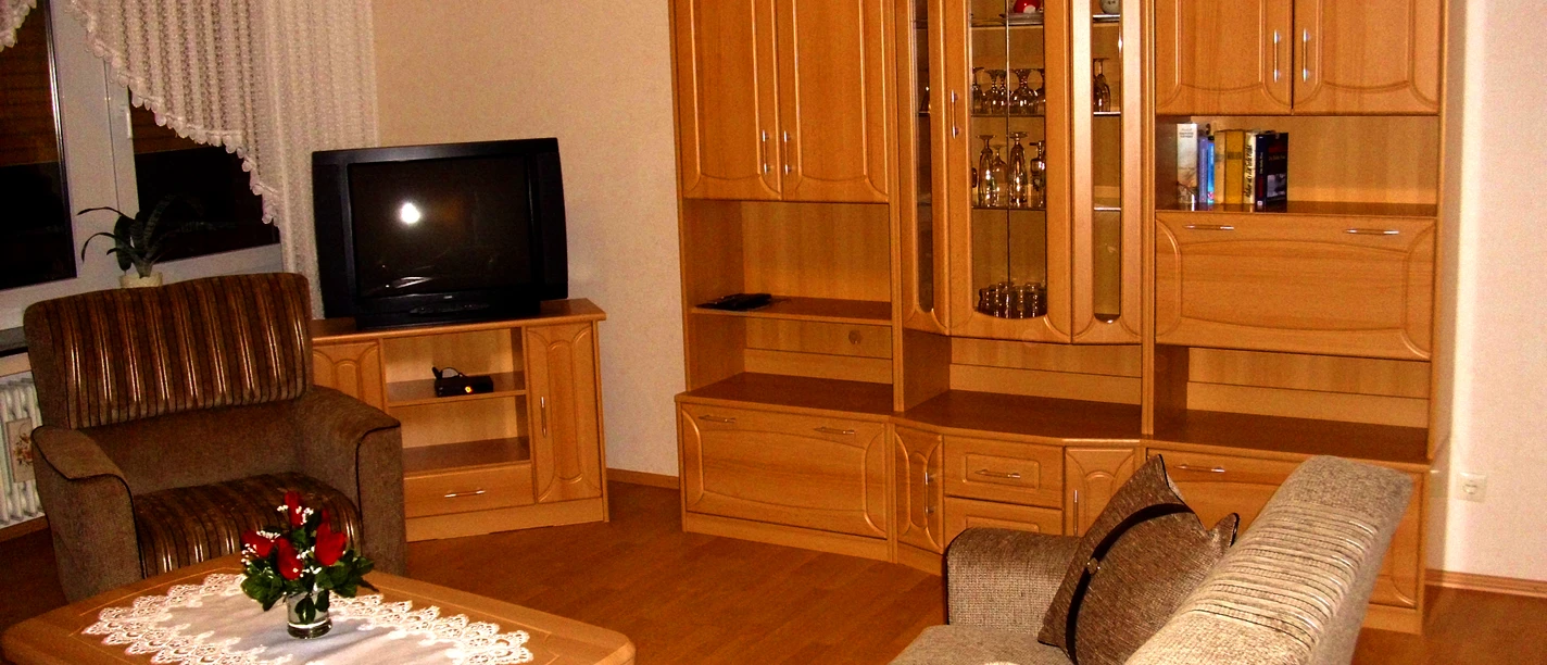 Wernekenschnieder.JPG Eine Wohnzimmer-Einrichtung mit Holzschränken, Sofa, Sessel, Fernseher und Couchtisch in warmen Tönen.