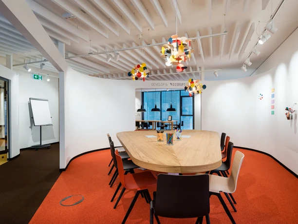 Denkfabrik im Innovation Center.jpg Moderner Konferenzraum mit ovalem Holztisch, bunten Stühlen und Designerleuchten auf rotem Teppich.</p>