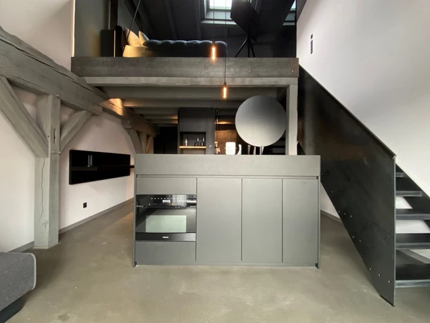 Ueberblick_Loft B+.jpg Moderne, graue Loftwohnung mit offener Küche, sichtbaren Holzbalken und Treppe zur offenen Empore.