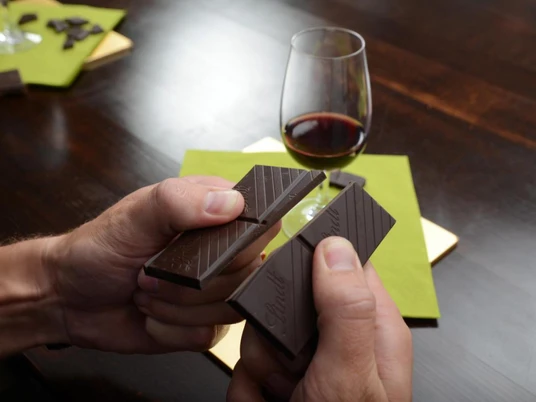 Verkostung Wein & Schokolade 2 Person bricht dunkle Schokolade, daneben ein Glas Rotwein auf einem grünen Untersetzer.</p>Person breaks dark chocolate, next to it a glass of red wine on a green coaster.</p> <p