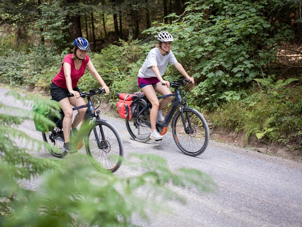 Zwei lachende Radfahrerinnen auf Radweg im Wald.
