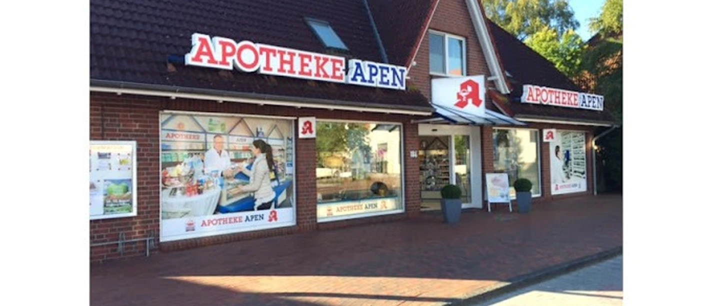 Apotheke-Apen_2023.jpg Das Bild zeigt die Frontansicht der Apotheke Apen mit einem markanten roten Schriftzug auf dem Dach. Die Fassade ist aus roten Backsteinen und verfügt über große Fenster, die Einblicke ins Innere gewähren. Vor der Apotheke befindet sich ein breiter Gehweg mit Pflastersteinen, begleitet von grünen Pflanzen in Töpfen. Die Apotheke strahlt eine einladende Atmosphäre aus.
