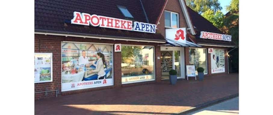 Apotheke-Apen_2023.jpg Das Bild zeigt die Frontansicht der Apotheke Apen mit einem markanten roten Schriftzug auf dem Dach. Die Fassade ist aus roten Backsteinen und verfügt über große Fenster, die Einblicke ins Innere gewähren. Vor der Apotheke befindet sich ein breiter Gehweg mit Pflastersteinen, begleitet von grünen Pflanzen in Töpfen. Die Apotheke strahlt eine einladende Atmosphäre aus.