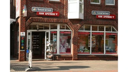 Fehn-Apotheke_Augustfehn.jpg Fehn-Apotheke mit rotem Klinkerbau, großen Fenstern und Eingangstür, umrahmt von Werbeschildern.