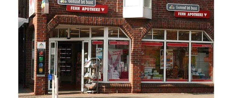 Fehn-Apotheke_Augustfehn.jpg Fehn-Apotheke mit rotem Klinkerbau, großen Fenstern und Eingangstür, umrahmt von Werbeschildern.