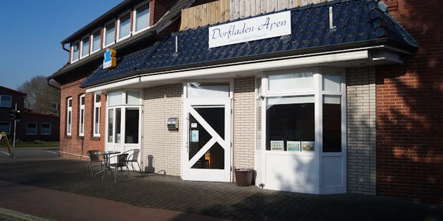 Dorfladen-Apen_Apen_2022.jpg Dorfladen in Apen mit roten Backsteinwänden, weißem Eingang und Sitzgelegenheiten im Freien.