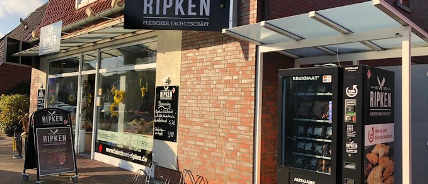 Fleischerei-Ripken_Augustfehn_2020.jpg Das Bild zeigt das Fleischerei Fachgeschäft Ripken, ein Backsteingebäude, in dem unten die Fleischeri ist. Vor dem Gebäude steht ein Automat, der Grillfleisch und Salate enthält