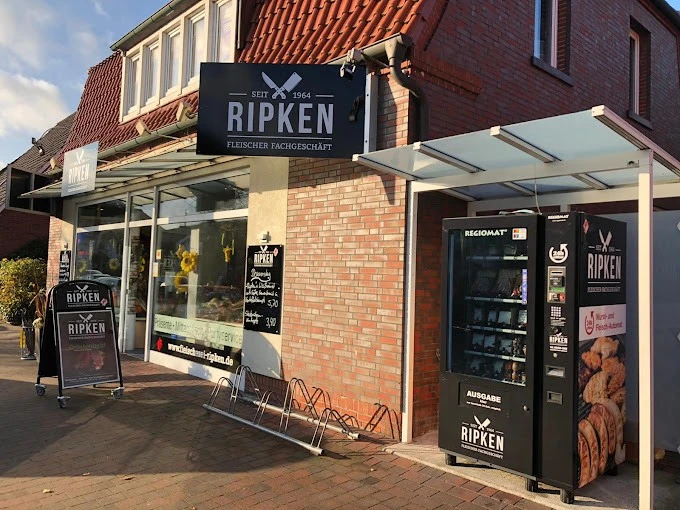 Fleischerei-Ripken_Augustfehn_2020.jpg Das Bild zeigt das Fleischerei Fachgeschäft Ripken, ein Backsteingebäude, in dem unten die Fleischeri ist. Vor dem Gebäude steht ein Automat, der Grillfleisch und Salate enthält