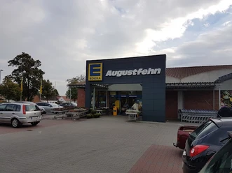 Edeka Augustfehn.jpg Das Bild zeigt den Eingangsbereich des Edeka-Markts in Augustfehn mit Kundenparkplatz und Einkaufswagen bei bewölktem Himmel.