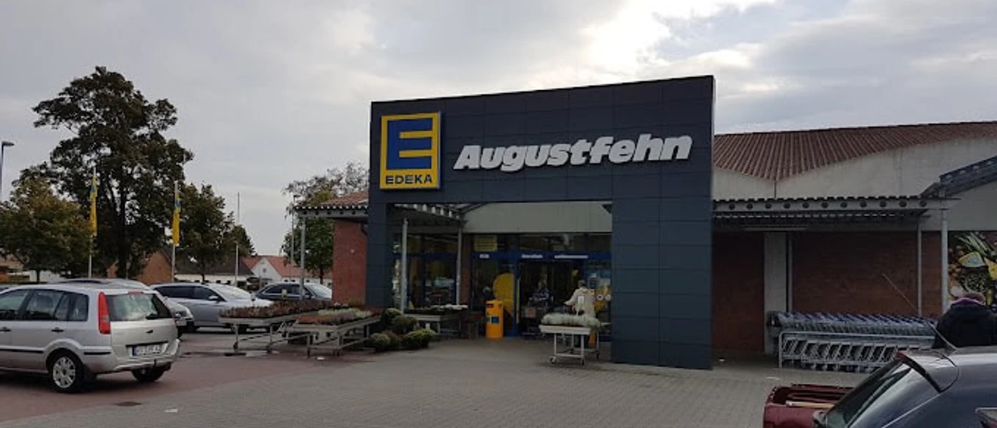 Edeka Augustfehn.jpg Das Bild zeigt den Eingangsbereich des Edeka-Markts in Augustfehn mit Kundenparkplatz und Einkaufswagen bei bewölktem Himmel.