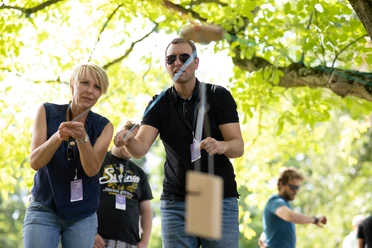 Teambuilding.jpg Teilnehmer werfen Holzstäbe bei einer freundlichen Outdoor-Veranstaltung unter einem Baum.