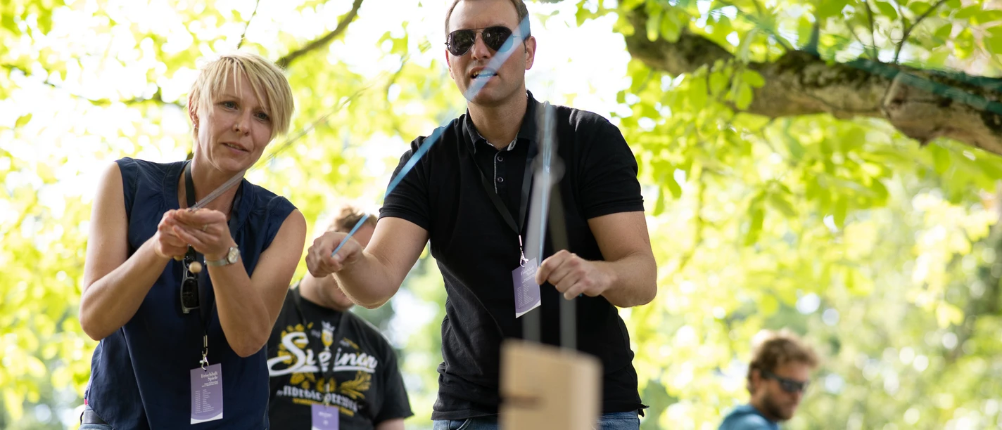 Teambuilding.jpg Teilnehmer werfen Holzstäbe bei einer freundlichen Outdoor-Veranstaltung unter einem Baum.