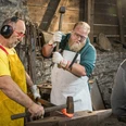 Werkzeugschmieden Zwei Männer bearbeiten glühendes Metall auf einem Amboss in einer Schmiedewerkstatt. Ein Zuschauer beobachtet.