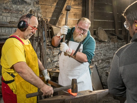Werkzeugschmieden Zwei Männer bearbeiten glühendes Metall auf einem Amboss in einer Schmiedewerkstatt. Ein Zuschauer beobachtet.
