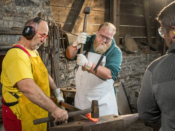 Werkzeugschmieden Zwei Männer bearbeiten glühendes Metall auf einem Amboss in einer Schmiedewerkstatt. Ein Zuschauer beobachtet.