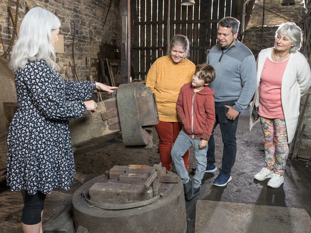 der letzte Hammer Eine Gruppe von Personen hört einer Dame in einem traditionellen Steinhaus mit alten Geräten zu.