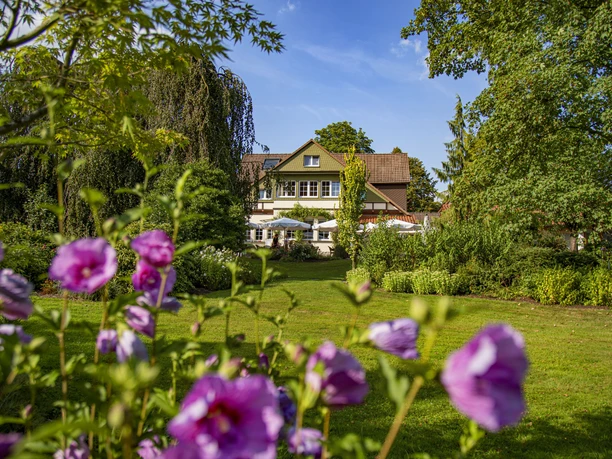 Ein weißes Landhaus mit roten Ziegeln und blühenden Blumen im Vordergrund, umgeben von Bäumen.