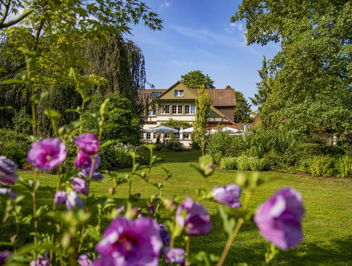 Ein weißes Landhaus mit roten Ziegeln und blühenden Blumen im Vordergrund, umgeben von Bäumen.