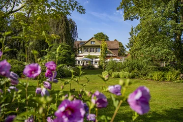 Haus Berkenkamp-Sommer.jpg Ein weißes Landhaus mit roten Ziegeln und blühenden Blumen im Vordergrund, umgeben von Bäumen.