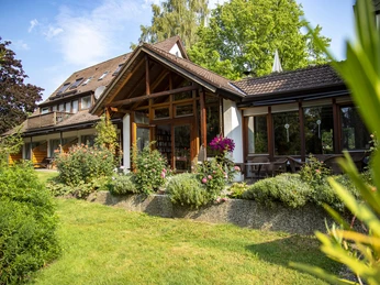 Schönes, modernes Haus mit großer Terrasse, blühenden Pflanzen und üppiger Umgebung im Grünen.