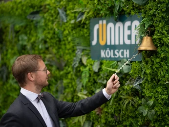 Kölsch Wall Kostümierter Mann läutet goldene Glocke vor grüner Pflanzenwand mit Werbeschild für Sünner Kölsch.Costumed man rings a golden bell in front of a green plant wall with an advertising sign for Sünner Kölsch.