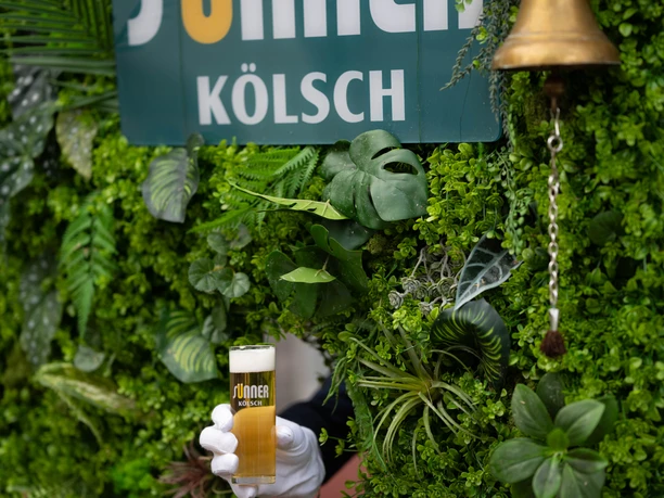 Kölsch Wall Green plant wall with "Sünner Kölsch" sign, next to it a bell and a hand holding a beer glass.</p> <p