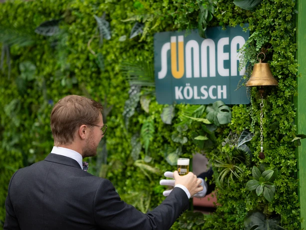 brauwelt-koeln-brauhaus-hochzeit-20.jpg Man in a suit drinking Kölsch in front of a green wall with a SUNNER sign and a bell.