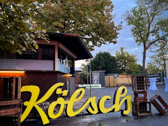 IMG_2669.jpg Gelbes "Kölsch"-Schild vor einem Holzhäuschen, umgeben von Bäumen auf nassem Asphalt.</p>Yellow "Kölsch" sign in front of a wooden house, surrounded by trees on wet asphalt.</p> <p