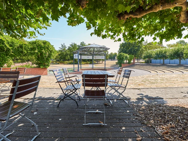Ringlokschuppen, Mülheim an der Ruhr <p>Ein Park mit leeren, hölzernen Bistrotischen und Stühlen unter grünen Bäumen, nahe einer Freilichtbühne.</p>