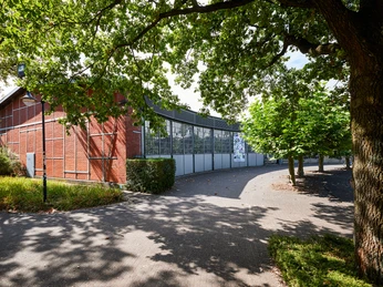 Ringlokschuppen Ruhr, Mülheim an der Ruhr <p>Rotes Backsteingebäude mit großflächigen Fenstern, umgeben von schattenspendenden Bäumen.</p>