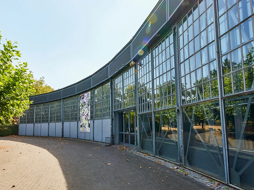 Ringlokschuppen Ruhr, Mülheim an der Ruhr Halbrundes Gebäude mit großflächigen Glasfassaden, Bäume und Sonnenstrahlen im Hintergrund.