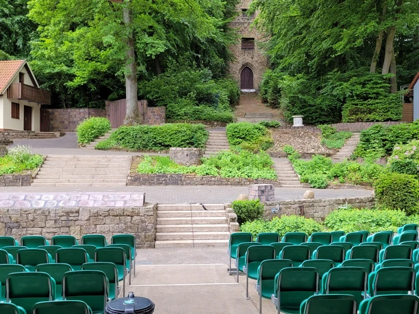 Grüne Sitzreihen umgeben von Bäumen und historischen Gebäuden, eine Freilichtbühne im Freien.