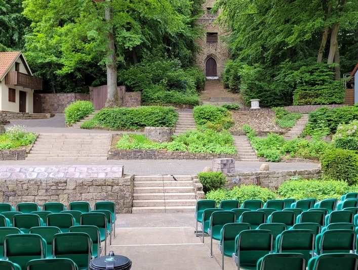 Grüne Sitzreihen umgeben von Bäumen und historischen Gebäuden, eine Freilichtbühne im Freien.