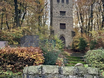 Steinerner Wehrturm umgeben von Herbstlaub, im Vordergrund ein altes Mauerstück mit Metallring.