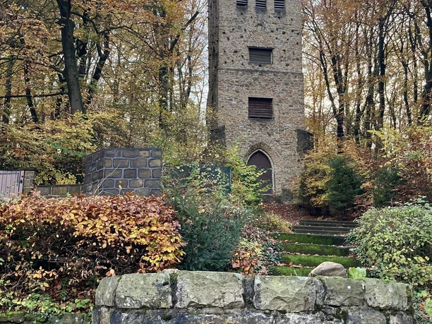 Steinerner Wehrturm umgeben von Herbstlaub, im Vordergrund ein altes Mauerstück mit Metallring.