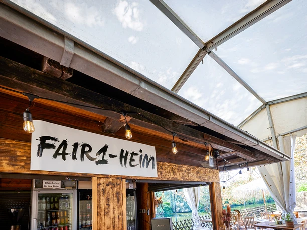<p>Im Bild ist ein Café unter einem Zeltpavillon zu sehen, mit einem Schild "FAIR1-HEIM".</p>