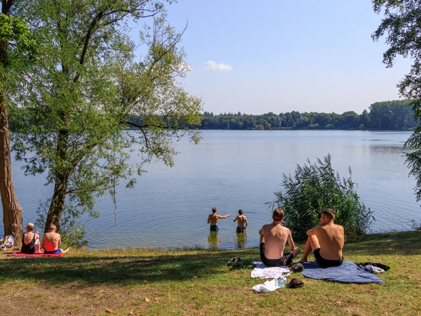 ostercappeln-kronensee©tol-foto-jurjen-drenth (7).JPG Menschen genießen einen Sommertag am schattigen Seeufer mit Bäumen und ruhigem Wasser.